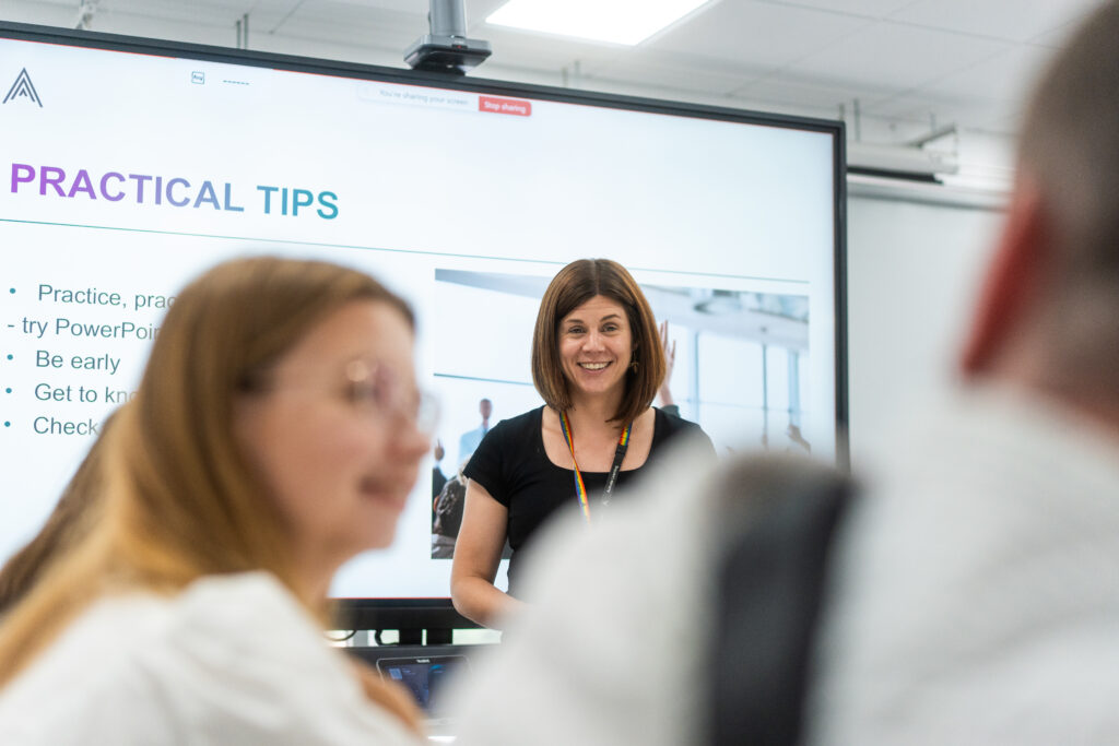 A female teacher wearing a lanyard standing in front of a screen presenting to a class